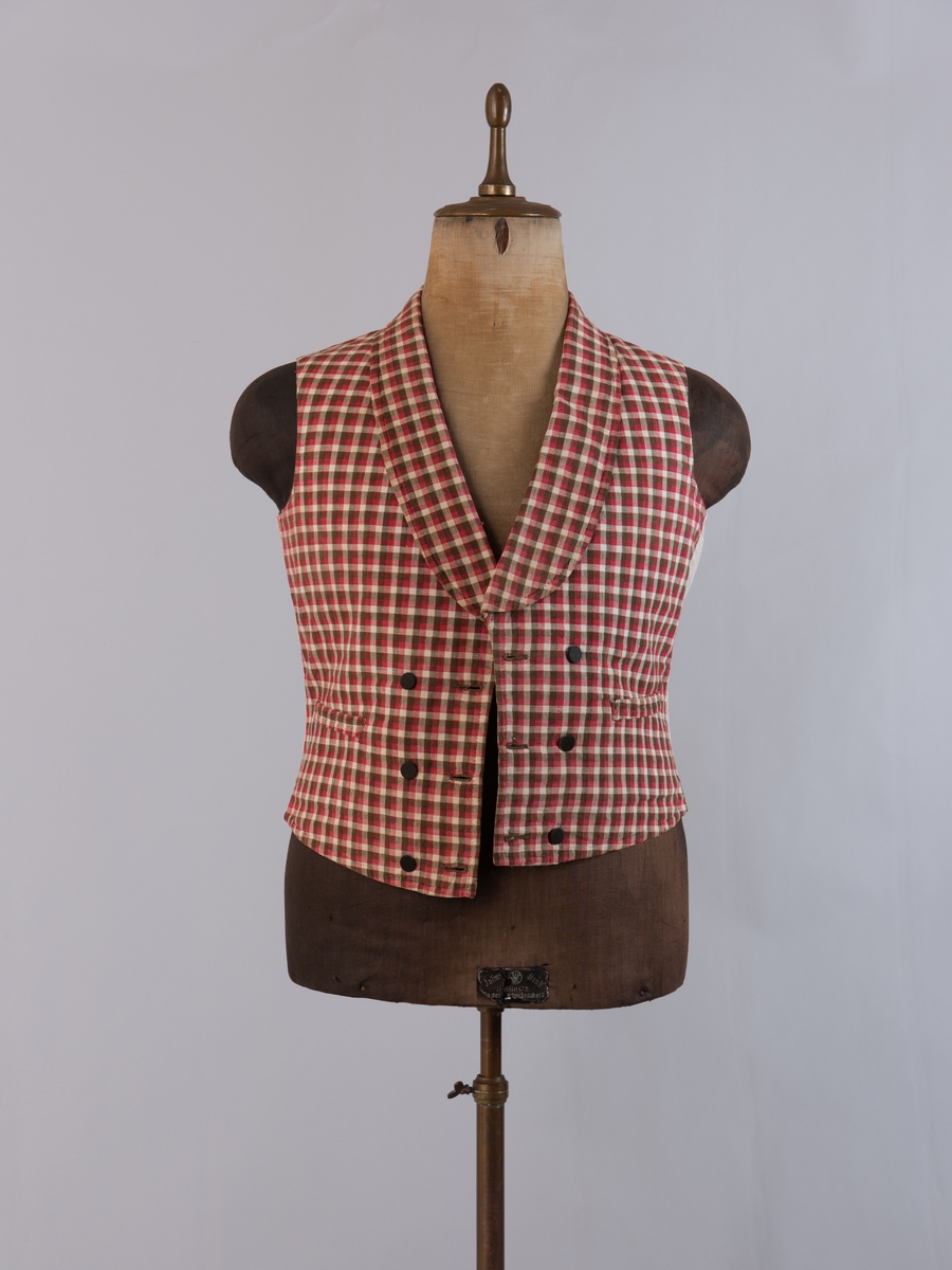 Vest i rutet bomullslerret i fargene rødt, olivengrønt/grønnnbrunt og ubleiket hvitt. Fôr og rygg i linlerret. Vesten er dobbeltspent med svarte, stofftrukne knapper. Den har sjalskrage og spensel i ryggen. Plagget er helt håndsydd. På forstykkene er det to lett skråstilte lommer og vesten buer ned midt foran.