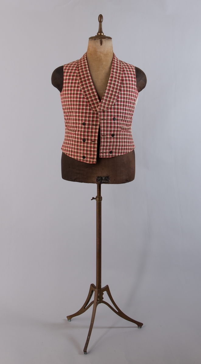 Vest i rutet bomullslerret i fargene rødt, olivengrønt/grønnnbrunt og ubleiket hvitt. Fôr og rygg i linlerret. Vesten er dobbeltspent med svarte, stofftrukne knapper. Den har sjalskrage og spensel i ryggen. Plagget er helt håndsydd. På forstykkene er det to lett skråstilte lommer og vesten buer ned midt foran.