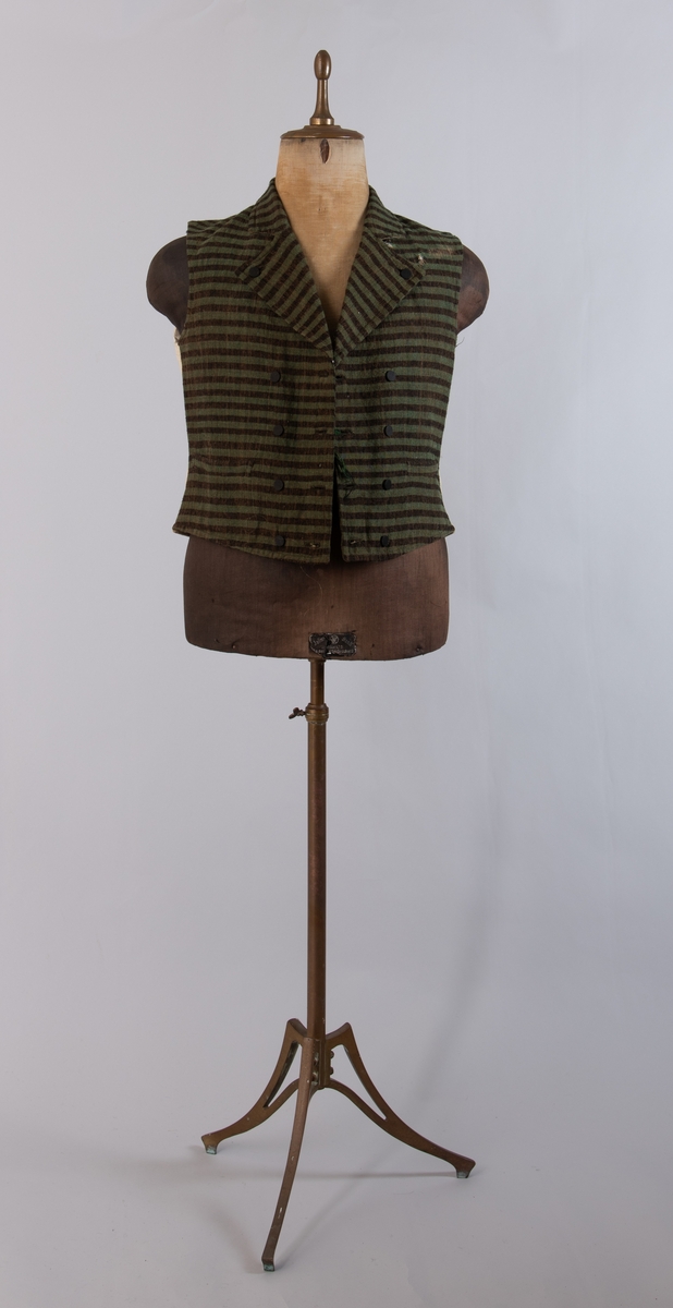 Dobbeltspent vest i ullverken, stripet i grønt og svart (sauesvart) med brun renning. Dobbel rygg i bomullslerret. Halvlinslerret (bomull/stry) i fôret i framstykkene. Vesten har fire knapphull sydd med kamgarnsullgarn og svarte knapper trukket med svart bomullssateng med brun renning. Plagget har nedbrettet krage og slag/reverser som kan kneppes mot bolen. Knapp og knapphull. Vesten buer ned midt framme og har svakt skråstilte lommer. Vattering av stry i framstykkene. I ryggen buer vesten opp midt bak. Den er vrangsydd langs ytterkantene og stukket ned med maskin, 0,8 cm fra kanten. (Registrering ved Bunad- og folkedraktrådet/Norsk institutt for bunad og folkedrakt v/Jon Fredrik Skauge, 2005, revidert v/Bjørn Sverre Hol Haugen, 2020.)