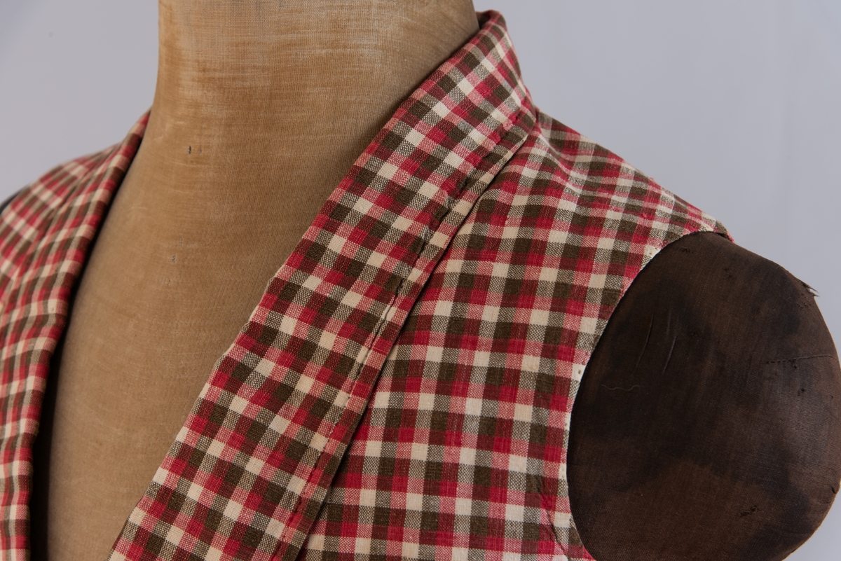 Vest i rutet bomullslerret i fargene rødt, olivengrønt/grønnnbrunt og ubleiket hvitt. Fôr og rygg i linlerret. Vesten er dobbeltspent med svarte, stofftrukne knapper. Den har sjalskrage og spensel i ryggen. Plagget er helt håndsydd. På forstykkene er det to lett skråstilte lommer og vesten buer ned midt foran.