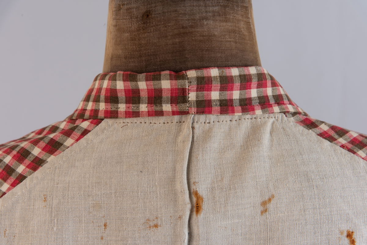 Vest i rutet bomullslerret i fargene rødt, olivengrønt/grønnnbrunt og ubleiket hvitt. Fôr og rygg i linlerret. Vesten er dobbeltspent med svarte, stofftrukne knapper. Den har sjalskrage og spensel i ryggen. Plagget er helt håndsydd. På forstykkene er det to lett skråstilte lommer og vesten buer ned midt foran.