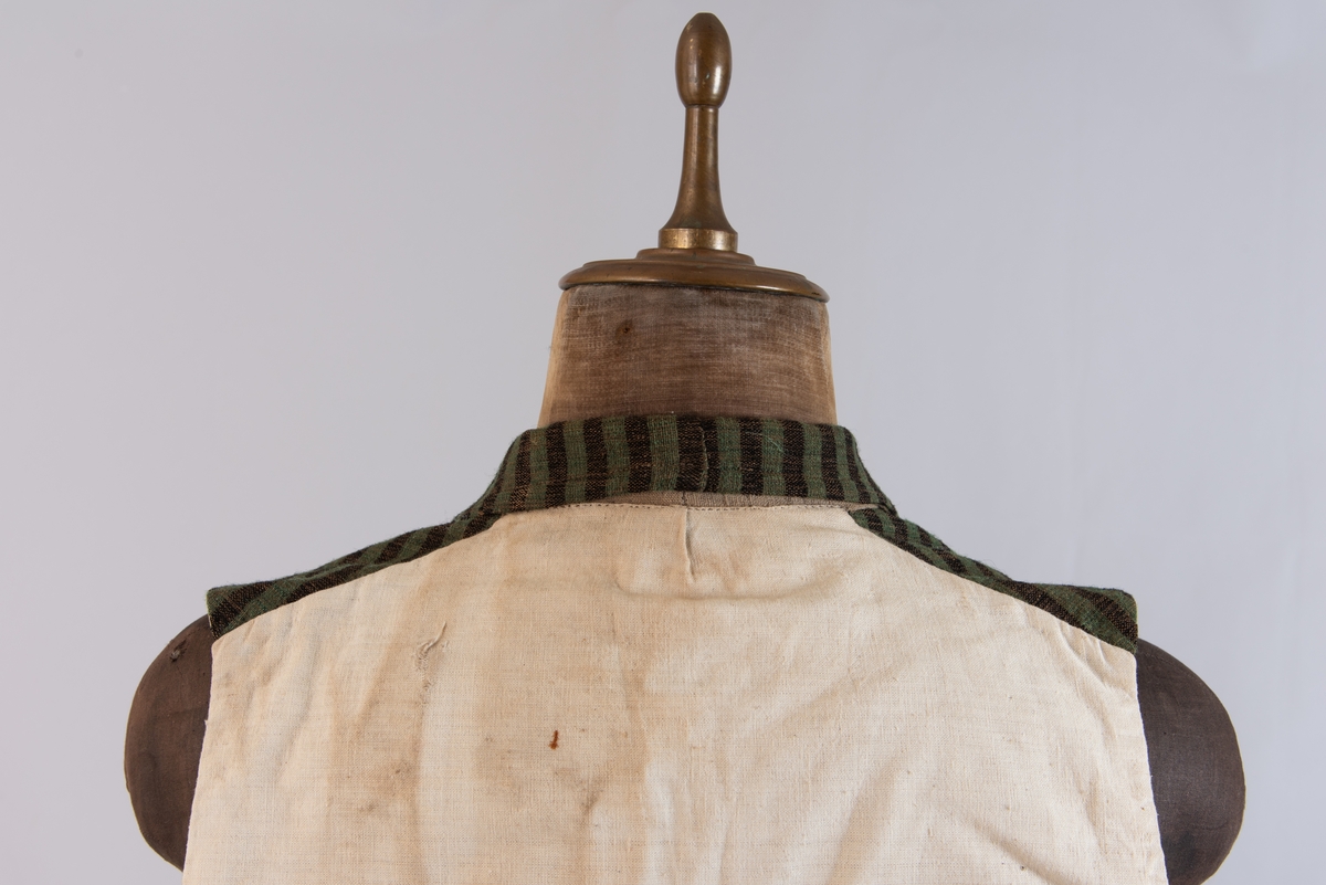 Dobbeltspent vest i ullverken, stripet i grønt og svart (sauesvart) med brun renning. Dobbel rygg i bomullslerret. Halvlinslerret (bomull/stry) i fôret i framstykkene. Vesten har fire knapphull sydd med kamgarnsullgarn og svarte knapper trukket med svart bomullssateng med brun renning. Plagget har nedbrettet krage og slag/reverser som kan kneppes mot bolen. Knapp og knapphull. Vesten buer ned midt framme og har svakt skråstilte lommer. Vattering av stry i framstykkene. I ryggen buer vesten opp midt bak. Den er vrangsydd langs ytterkantene og stukket ned med maskin, 0,8 cm fra kanten. (Registrering ved Bunad- og folkedraktrådet/Norsk institutt for bunad og folkedrakt v/Jon Fredrik Skauge, 2005, revidert v/Bjørn Sverre Hol Haugen, 2020.)