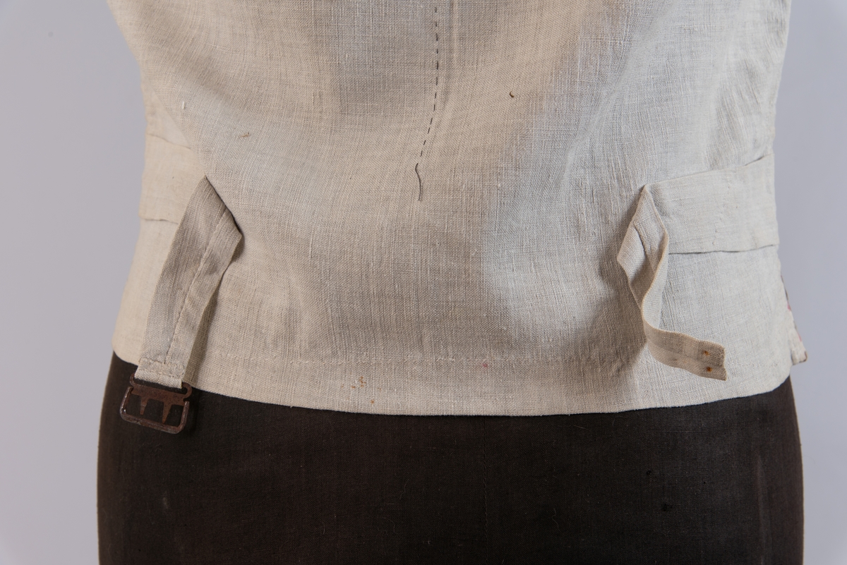 Vest i rutet bomullslerret i fargene rødt, olivengrønt/grønnnbrunt og ubleiket hvitt. Fôr og rygg i linlerret. Vesten er dobbeltspent med svarte, stofftrukne knapper. Den har sjalskrage og spensel i ryggen. Plagget er helt håndsydd. På forstykkene er det to lett skråstilte lommer og vesten buer ned midt foran.