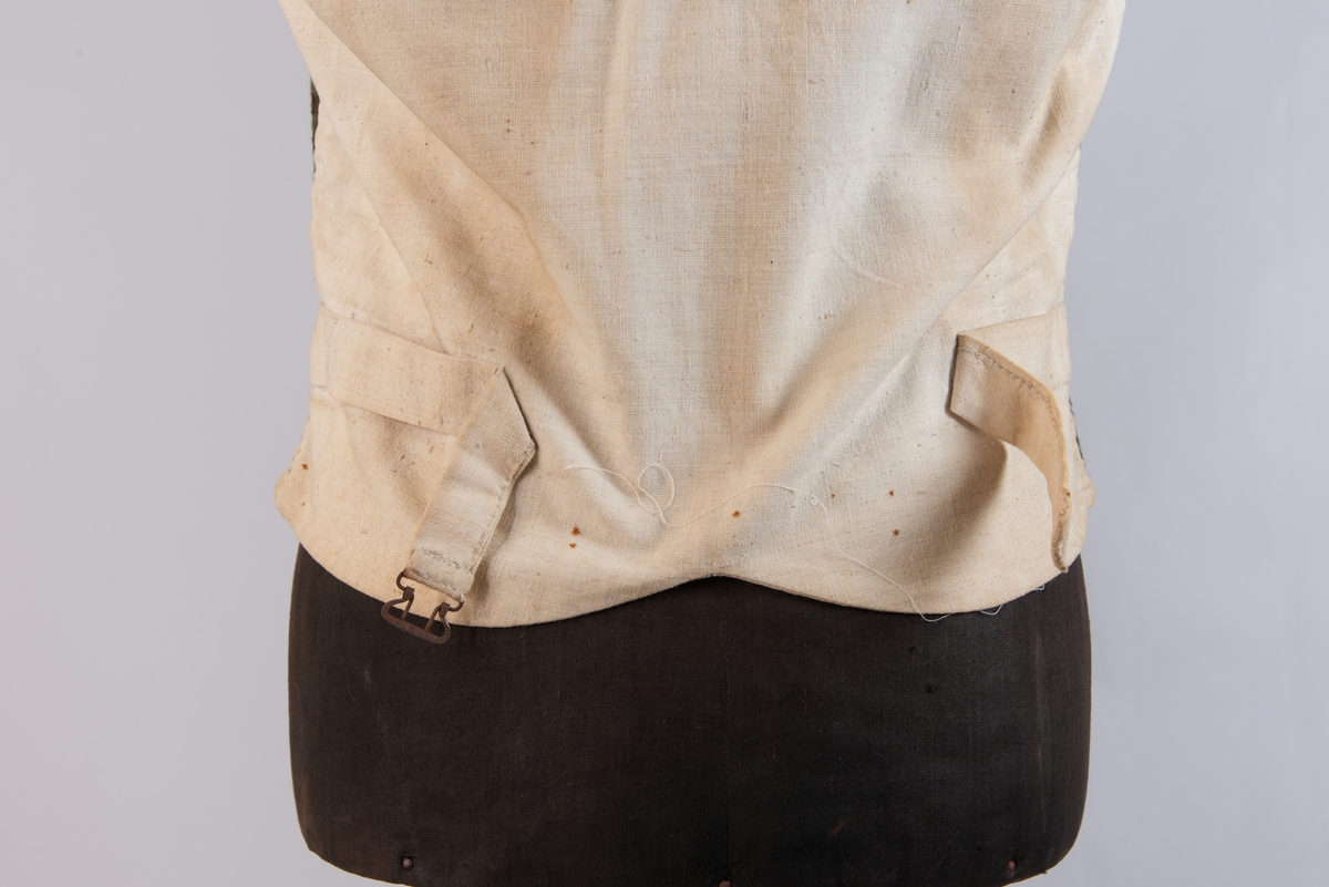 Dobbeltspent vest i ullverken, stripet i grønt og svart (sauesvart) med brun renning. Dobbel rygg i bomullslerret. Halvlinslerret (bomull/stry) i fôret i framstykkene. Vesten har fire knapphull sydd med kamgarnsullgarn og svarte knapper trukket med svart bomullssateng med brun renning. Plagget har nedbrettet krage og slag/reverser som kan kneppes mot bolen. Knapp og knapphull. Vesten buer ned midt framme og har svakt skråstilte lommer. Vattering av stry i framstykkene. I ryggen buer vesten opp midt bak. Den er vrangsydd langs ytterkantene og stukket ned med maskin, 0,8 cm fra kanten. (Registrering ved Bunad- og folkedraktrådet/Norsk institutt for bunad og folkedrakt v/Jon Fredrik Skauge, 2005, revidert v/Bjørn Sverre Hol Haugen, 2020.)