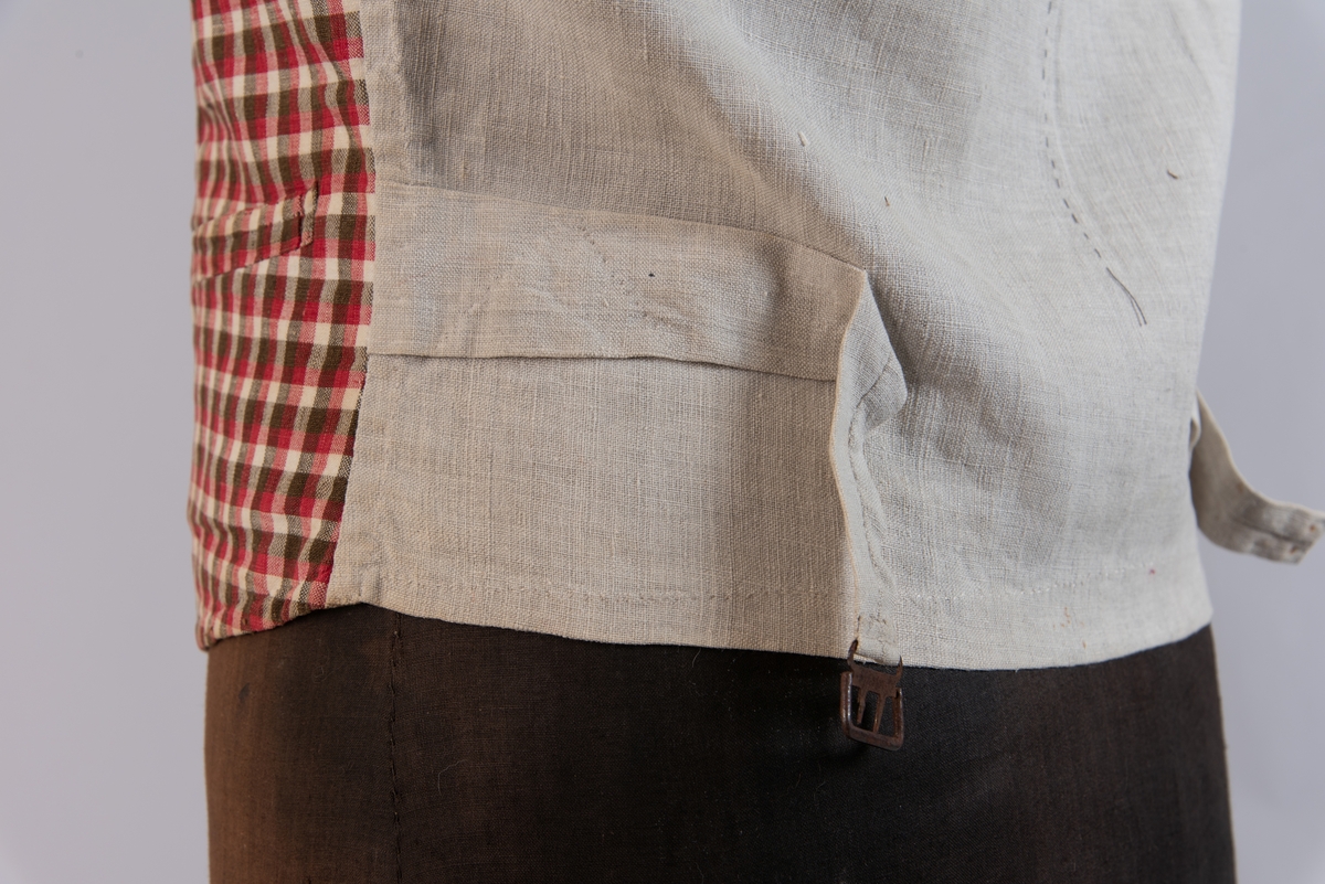 Vest i rutet bomullslerret i fargene rødt, olivengrønt/grønnnbrunt og ubleiket hvitt. Fôr og rygg i linlerret. Vesten er dobbeltspent med svarte, stofftrukne knapper. Den har sjalskrage og spensel i ryggen. Plagget er helt håndsydd. På forstykkene er det to lett skråstilte lommer og vesten buer ned midt foran.