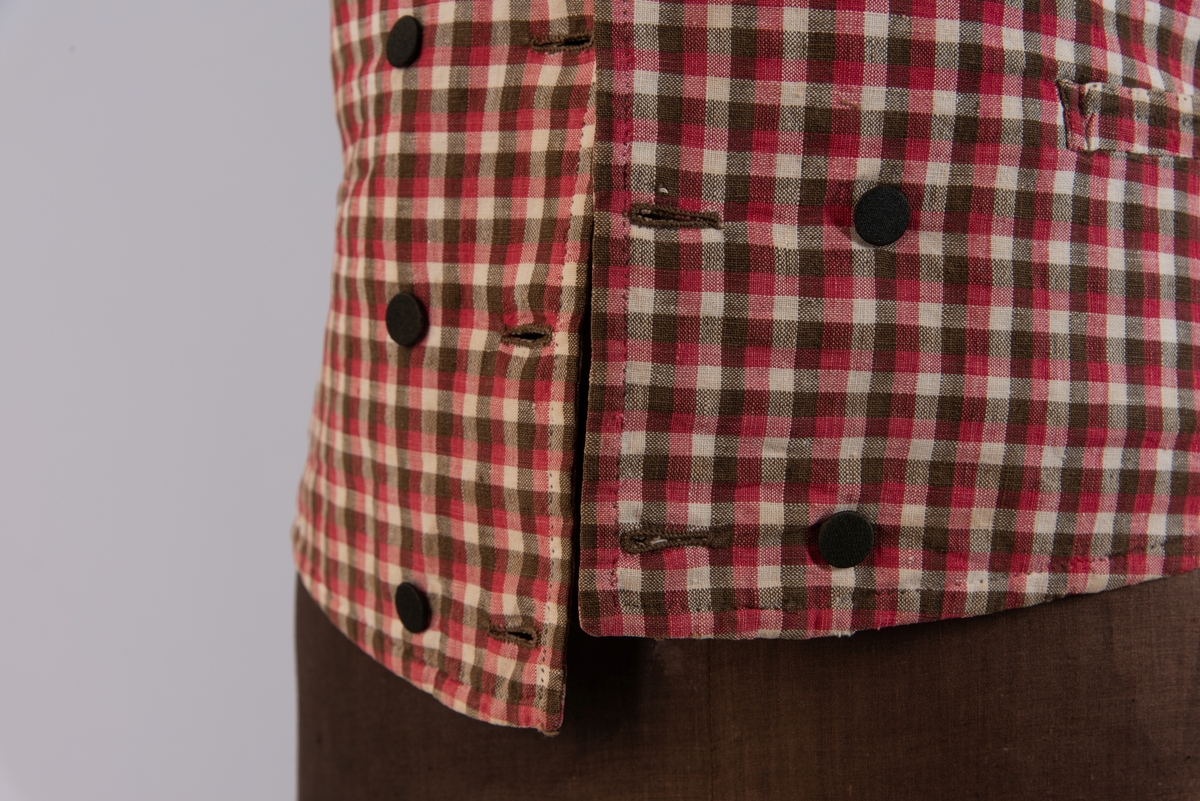 Vest i rutet bomullslerret i fargene rødt, olivengrønt/grønnnbrunt og ubleiket hvitt. Fôr og rygg i linlerret. Vesten er dobbeltspent med svarte, stofftrukne knapper. Den har sjalskrage og spensel i ryggen. Plagget er helt håndsydd. På forstykkene er det to lett skråstilte lommer og vesten buer ned midt foran.