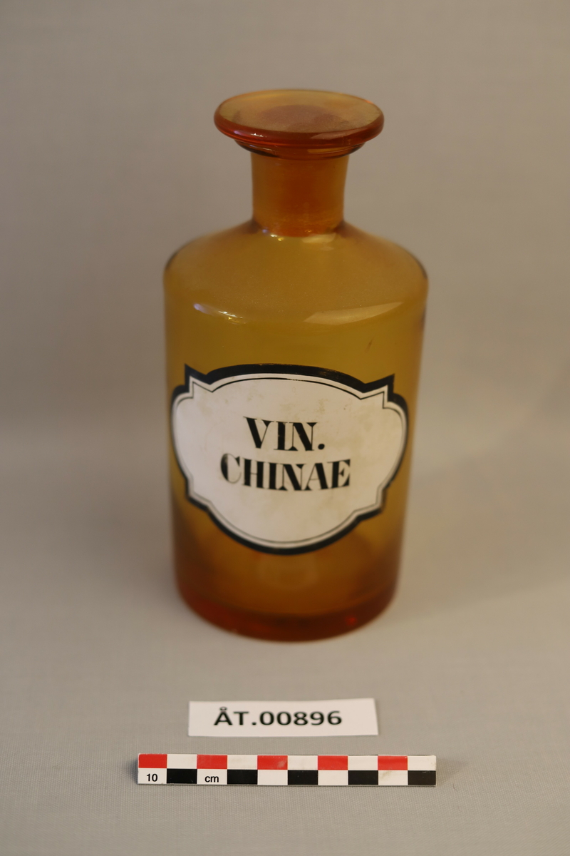 Brun, rund glassflaske/krukke med lok/kork. Kvit pålimt lapp på flaska med svart tekst: VIN.CHINAE
