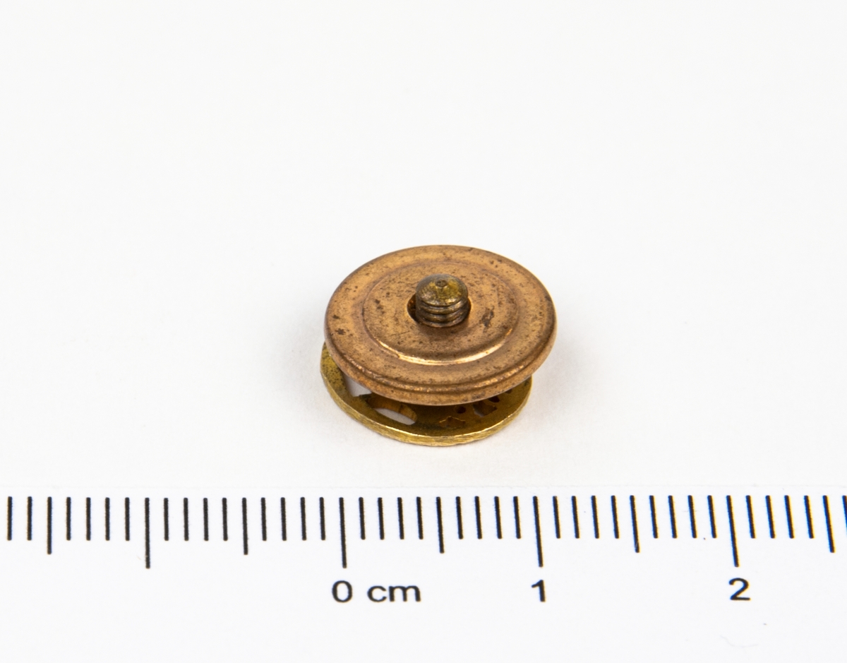 Märke med skruvfäste på baksidan. Motiv: ett genombrutet vikingaskepp och under skeppet en genombruten text "10V".