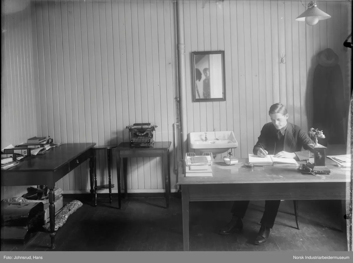 En mann som sitter ved en pult og skriver. På pulten står det en telefon. I speilet er fotografen synlig. Interiør fra kontor.