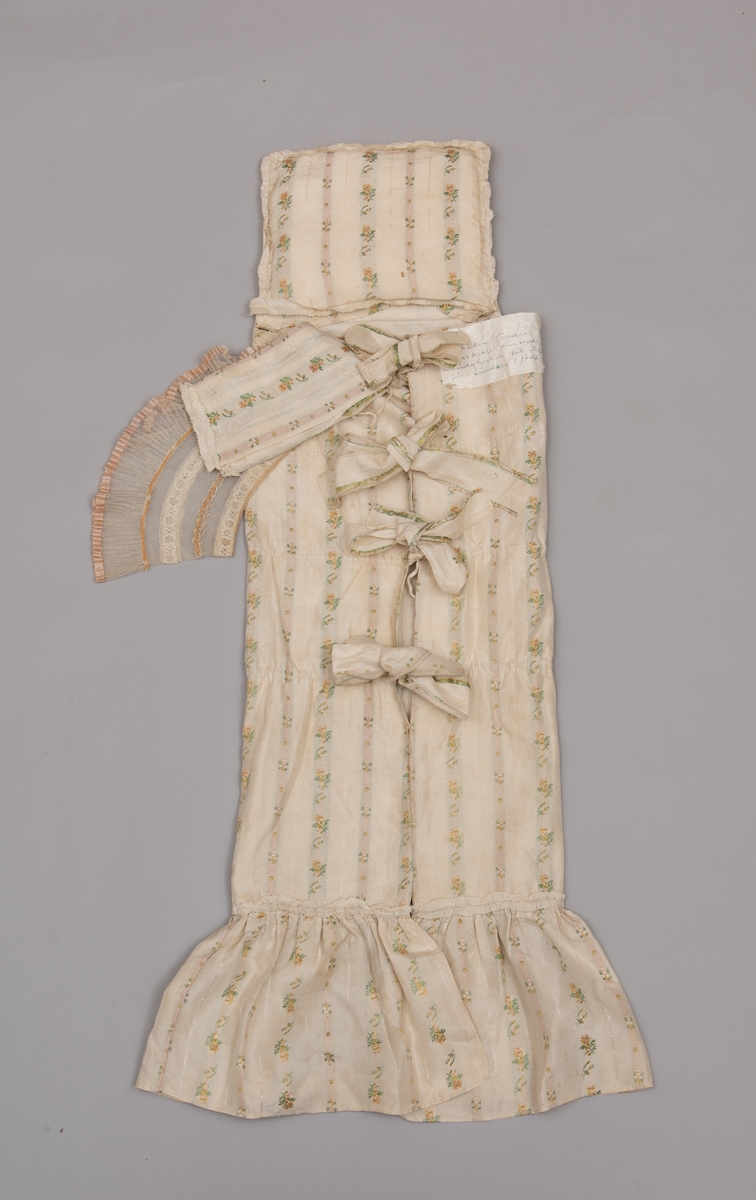 Dåpsløyert med pute samt løse ermer og en blondekrage. Løyerten er sydd av et ecrufarget silkestoff med stripemønster hvor det er innvevd blomster og blader. Løyerten lukkes med to sett knyteband, det ene settet er enkle bendelband som også snøres gjennom løpeganger, mens det andre er silkeband sydd av stoffet. Langs nederkant har løyerten en rynket kant av hovedstoffet samt et rynket dekorband av ensfarget, ecrufarget silke. Løyerten er fôret med linlerret. Ermene er sydd av hovedstoffet og har dekor av samme rynkede silkeband, men er uten fôr. Trolig har ermene vært tråklet til ei undertrøye som dåpsbarnet har hatt under reiven. Kragen, som er av tyllblonde, trolig i lin med silketråder og silkeband, er vid, så den har trolig vært rynket med ei snor og tråklet til undertrøya. Da barnet lå som en reivet bylt inne i løyerten, var det bare ermene som kom til syne, derfor var det ikke nødvendig med ei hel trøye. Alternativt kan det være at ermene tidligere har sittet på ei silketrøye som nå er fjernet.