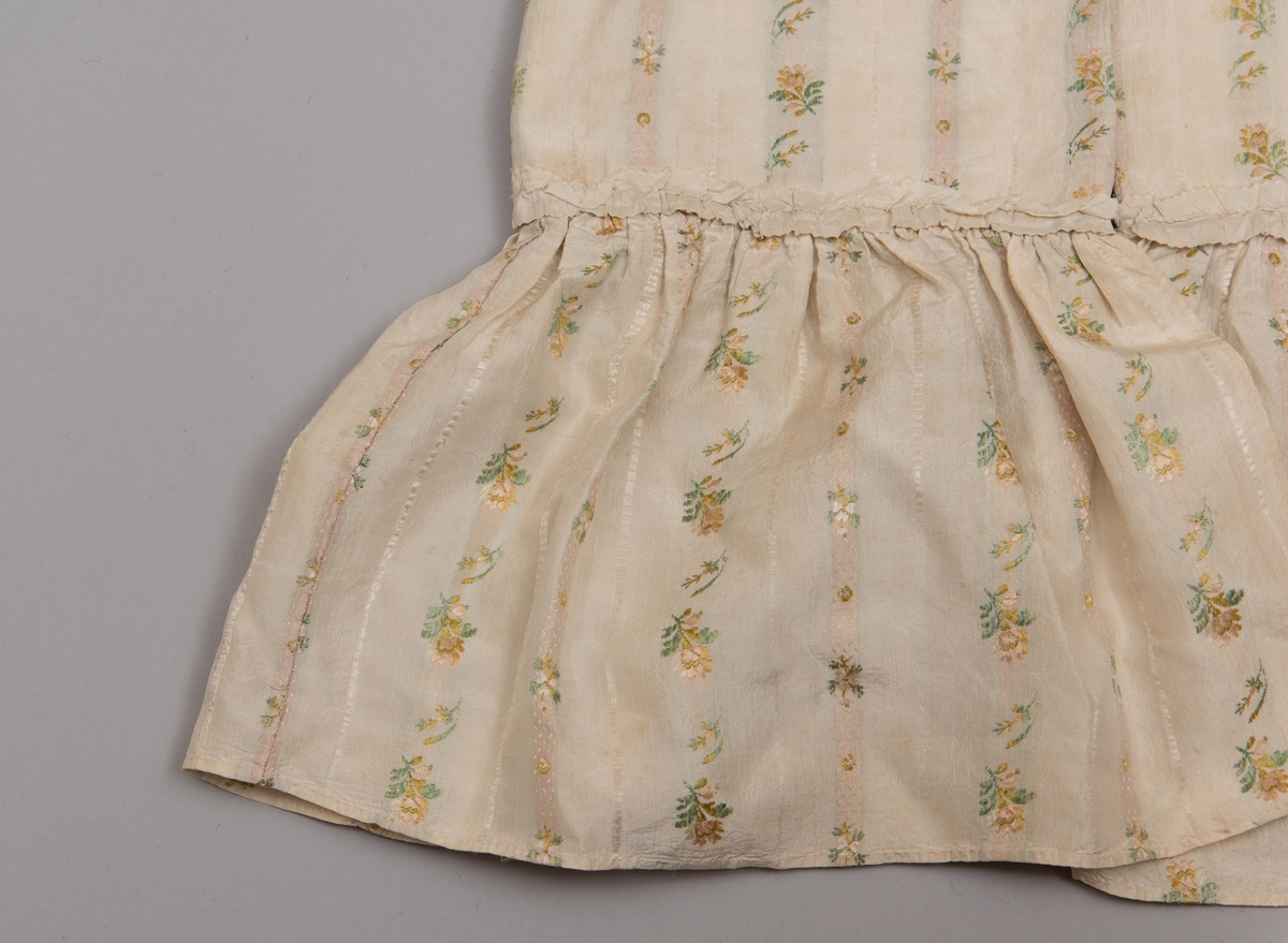 Dåpsløyert med pute samt løse ermer og en blondekrage. Løyerten er sydd av et ecrufarget silkestoff med stripemønster hvor det er innvevd blomster og blader. Løyerten lukkes med to sett knyteband, det ene settet er enkle bendelband som også snøres gjennom løpeganger, mens det andre er silkeband sydd av stoffet. Langs nederkant har løyerten en rynket kant av hovedstoffet samt et rynket dekorband av ensfarget, ecrufarget silke. Løyerten er fôret med linlerret. Ermene er sydd av hovedstoffet og har dekor av samme rynkede silkeband, men er uten fôr. Trolig har ermene vært tråklet til ei undertrøye som dåpsbarnet har hatt under reiven. Kragen, som er av tyllblonde, trolig i lin med silketråder og silkeband, er vid, så den har trolig vært rynket med ei snor og tråklet til undertrøya. Da barnet lå som en reivet bylt inne i løyerten, var det bare ermene som kom til syne, derfor var det ikke nødvendig med ei hel trøye. Alternativt kan det være at ermene tidligere har sittet på ei silketrøye som nå er fjernet.