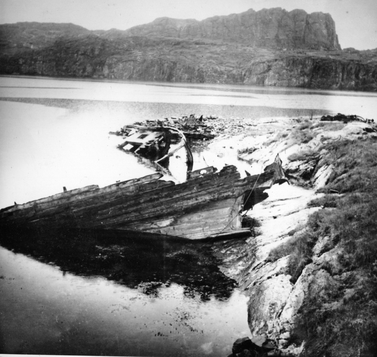 Telavåg, ruiner, forkulla fiskebåt i fjæra. Nipafjellet i bakgrunnen.