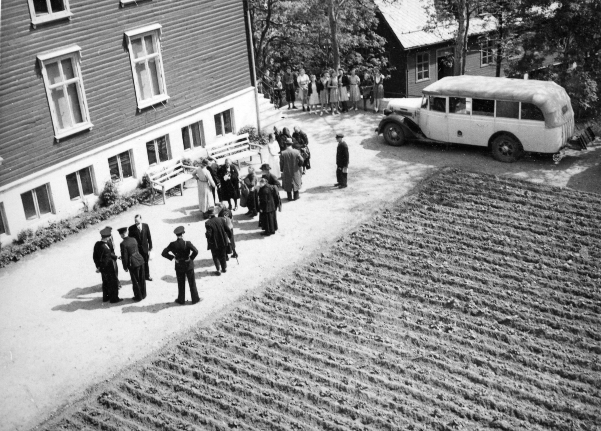 Bussen som har frakta dei eldre frå Storetveit til Framnes står parkert på skuleområdet. Eldre personar og politibetjentar utanfor skulebygningen. Ein flokk med elevar frå framhaldskuleklassen på Framnes står i bakgrunnen og ser.