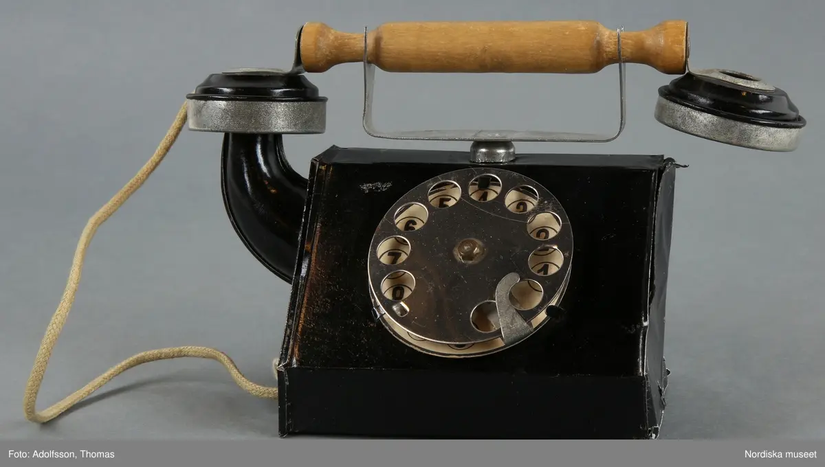 Inventering Sesam 1996-1999:
L  20   B  7,5   H  11,5 (cm)  
Telefon, leksak, av bockad och svartlackerad plåt, bordsmodell, rak form, klyka och nummerskiva av olackerad plåt, svarta siffror på vitt papper, lur med handtag av obehandlat lövträ, sladd av ljusgrön flätad tråd. När man vrider nummerskivan uppstår ett plingande ljud.
Har använts av Gunnar Hagelin, f 1925.
Anna Womack jan 1998