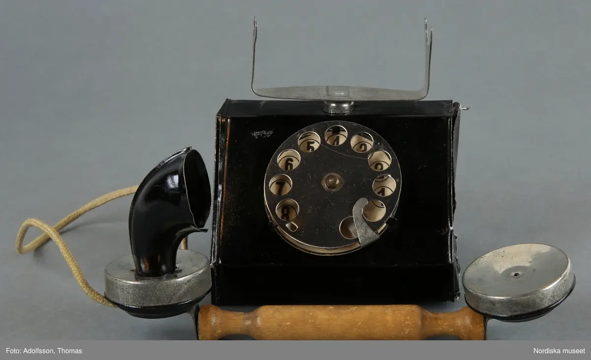 Inventering Sesam 1996-1999:
L  20   B  7,5   H  11,5 (cm)  
Telefon, leksak, av bockad och svartlackerad plåt, bordsmodell, rak form, klyka och nummerskiva av olackerad plåt, svarta siffror på vitt papper, lur med handtag av obehandlat lövträ, sladd av ljusgrön flätad tråd. När man vrider nummerskivan uppstår ett plingande ljud.
Har använts av Gunnar Hagelin, f 1925.
Anna Womack jan 1998