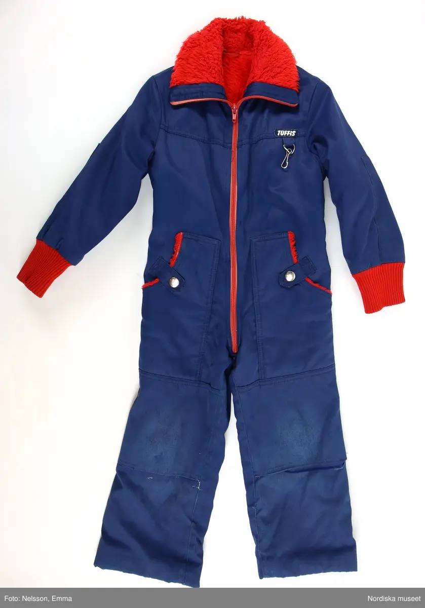 Katalogkort:
"Overall f. pojke av mörkblå s.k. "bäver-nylon" kypertvävnad av 50% polyamid och 50% bomull (mycket slitstark används till arbetskläder och barnkläder) fodrad med röd acrylpile i överdelen och med syntetvadd rutstickad med röd nylon i benen och ärmarna. 
Helskuren modell med besparing på axlarna fram, resår i midjan bak, isydd lång ärm med röda muddar av acryltrikå, lång rött blixtlås fram ned till grenen, rak krage med rött pilefoder, 2 påsydda framfickor med rött pilefoder och knäppta med slejf och nitad tryckknapp. Förstärkt med extra tyg på knäna. Fastsydd vitmetallhake på bröstet att fästa skidhandskarna i. Märke i vitt: Tuffis Reflexbricka i snöre fäst bak på kragen med säkerhetsnål."