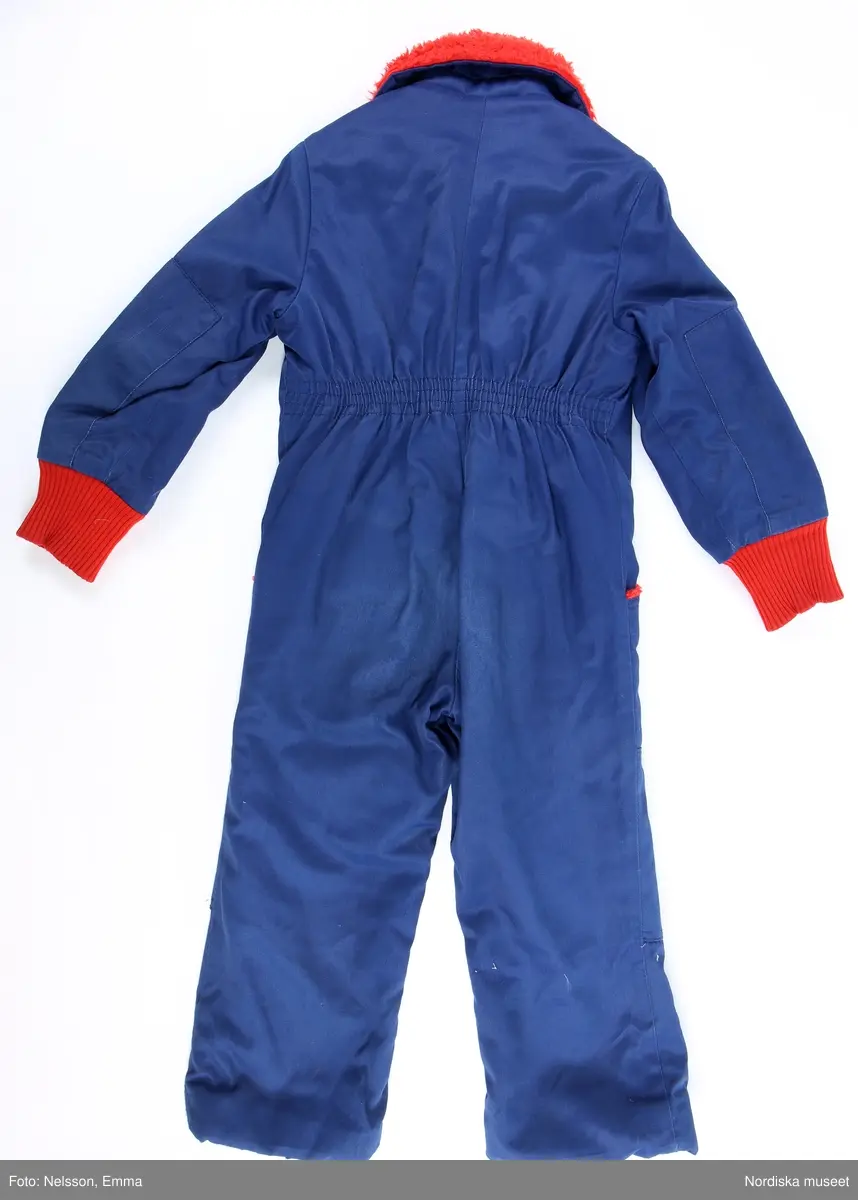 Katalogkort:
"Overall f. pojke av mörkblå s.k. "bäver-nylon" kypertvävnad av 50% polyamid och 50% bomull (mycket slitstark används till arbetskläder och barnkläder) fodrad med röd acrylpile i överdelen och med syntetvadd rutstickad med röd nylon i benen och ärmarna. 
Helskuren modell med besparing på axlarna fram, resår i midjan bak, isydd lång ärm med röda muddar av acryltrikå, lång rött blixtlås fram ned till grenen, rak krage med rött pilefoder, 2 påsydda framfickor med rött pilefoder och knäppta med slejf och nitad tryckknapp. Förstärkt med extra tyg på knäna. Fastsydd vitmetallhake på bröstet att fästa skidhandskarna i. Märke i vitt: Tuffis Reflexbricka i snöre fäst bak på kragen med säkerhetsnål."
