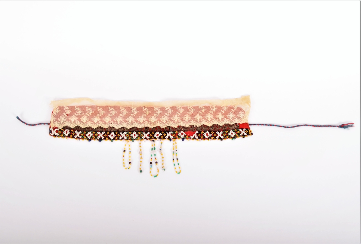 Lad, pannelin (a i raudt toskaftvove vadmel, påsett ein "perlebendel" av kvite, gule, grønne og svart perler. Perlesnor langst ytterste kanten av gule og blanke små perler, og med større grønne, blåe, brune og blanke perler med ca 2 cm mellomrom. Midt bak i nakken fem perleheng men blanke, gullblanke, blå, gule og svarte perler. 
Innanfor perlebendelen eit band i sølvagraman. 
Sekundert påsette ein kvit tyllblonde (b) (Ref. registreringar hjå BRF i 1978, 1981, 1988)
I kvar ende ei slindre snor