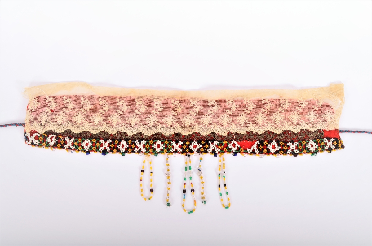 Lad, pannelin (a i raudt toskaftvove vadmel, påsett ein "perlebendel" av kvite, gule, grønne og svart perler. Perlesnor langst ytterste kanten av gule og blanke små perler, og med større grønne, blåe, brune og blanke perler med ca 2 cm mellomrom. Midt bak i nakken fem perleheng men blanke, gullblanke, blå, gule og svarte perler. 
Innanfor perlebendelen eit band i sølvagraman. 
Sekundert påsette ein kvit tyllblonde (b) (Ref. registreringar hjå BRF i 1978, 1981, 1988)
I kvar ende ei slindre snor
