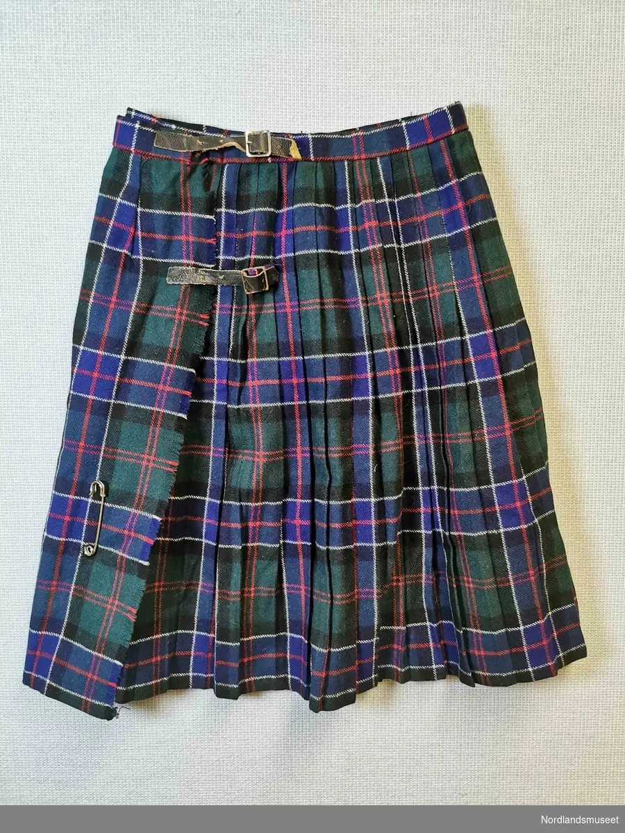 Rutete skjørt/kilt i blåttt, grønt, rødt og hvitt. Folder. Lukking med to lærhemper. En stor sikkerhetsnål er festet på venstre side.