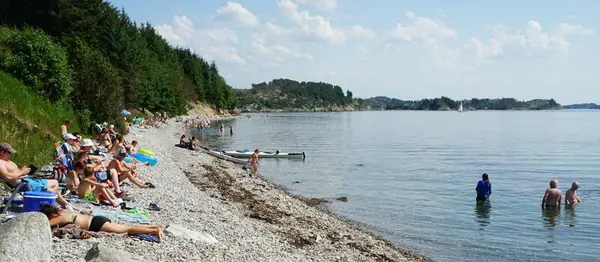Folk ligg i badetøy på rullesteinstranda på vestsida av Herdla