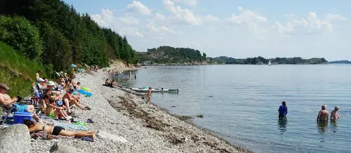 Folk ligg i badetøy på rullesteinstranda på vestsida av Herdla