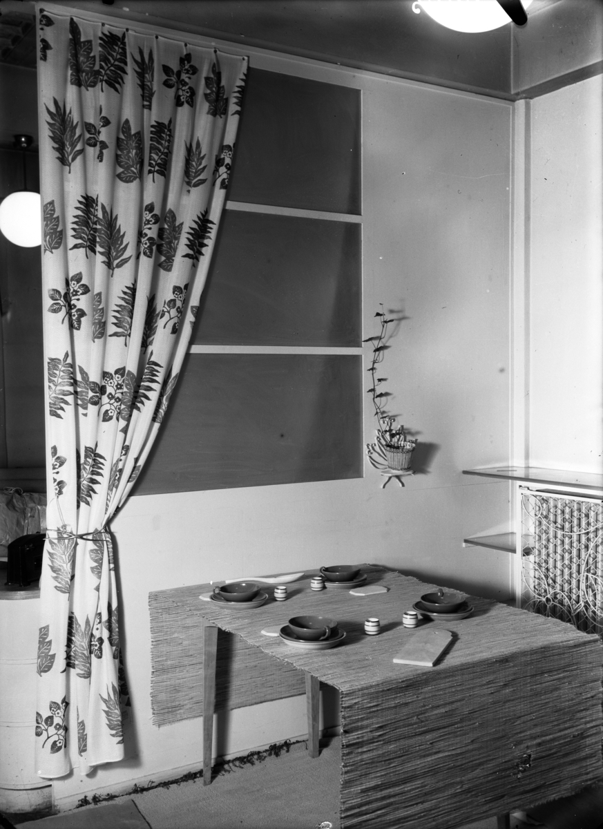 Trykt tekstil med blomstermotiv, drapert som gardin på vegg, foran fiktivt vindu, på andre siden av vinduet en potteplante, foran vinduet et frokostbord dekket med store tekopper og tallerkener i keramikk, eggeglass og små skjærefjøler til alle fire kuverter, på grovt vevd duk. I bakgrunnen en radiator. Publisert i Bonytt, trolig 1949-55.
