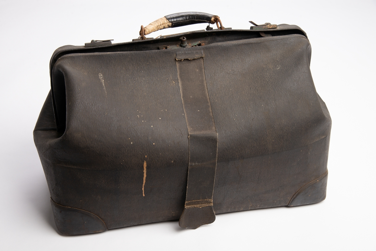 Svart koffert kunstskinn med teksturert overflate. Rektangulær bunn med plastforsterkninger i hjørnene. Fem metall knotter under. Lukkning i toppen via låsbar metallbøyle med plastbelagt hank, flapp i plast til å feste i låsen. Ett stort innvendig rom med en smal stikklomme på hver langside.