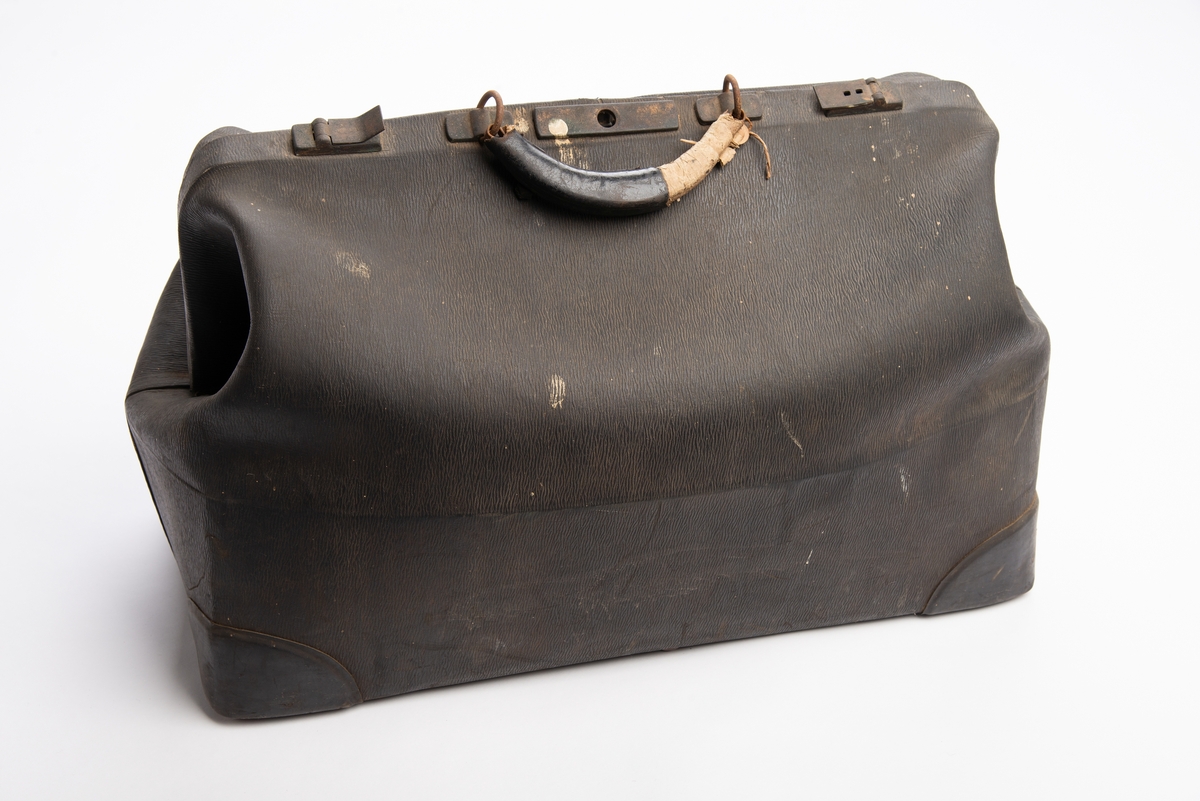 Svart koffert kunstskinn med teksturert overflate. Rektangulær bunn med plastforsterkninger i hjørnene. Fem metall knotter under. Lukkning i toppen via låsbar metallbøyle med plastbelagt hank, flapp i plast til å feste i låsen. Ett stort innvendig rom med en smal stikklomme på hver langside.