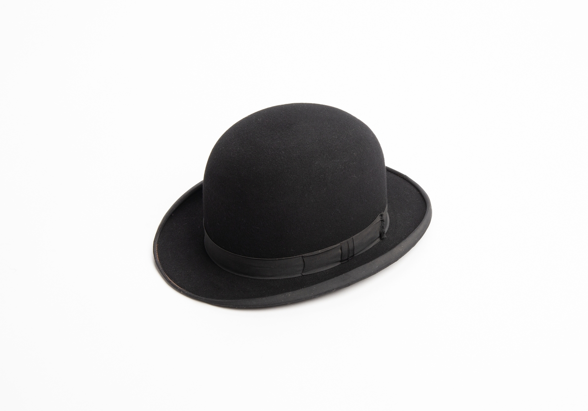 Svart bowlerhatt med svakt oppoverbuet ytterkant på brem. Rund pull og oval brem. Svart bånd med sløyfe i overgangen mellom brem og pull. Svart bånd langs ytterkanten av bremmen. Bremmen er stivet opp med metalltråd i ytterkant.. Svettebånd av lær på innsiden. Mellom svettebånd og pull ligger det avispapir.