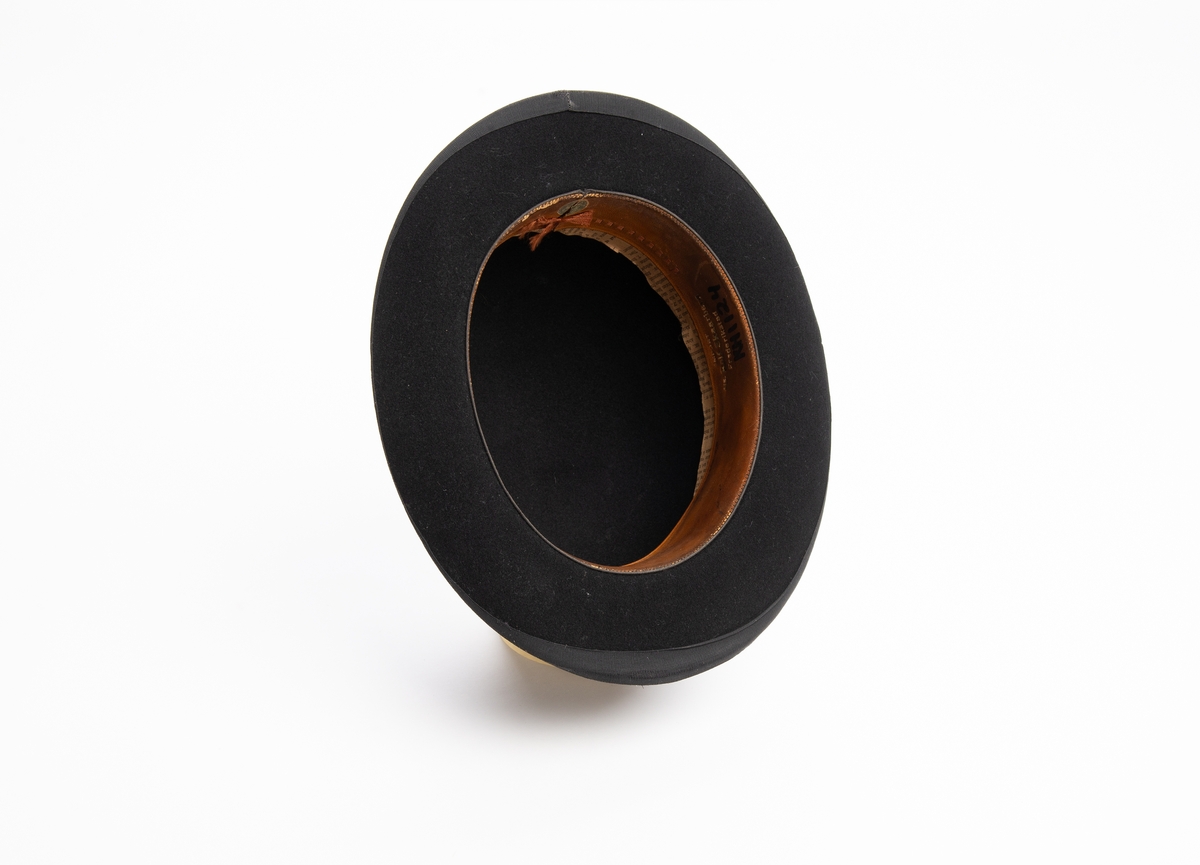 Svart bowlerhatt med svakt oppoverbuet ytterkant på brem. Rund pull og oval brem. Svart bånd med sløyfe i overgangen mellom brem og pull. Svart bånd langs ytterkanten av bremmen. Bremmen er stivet opp med metalltråd i ytterkant.. Svettebånd av lær på innsiden. Mellom svettebånd og pull ligger det avispapir.