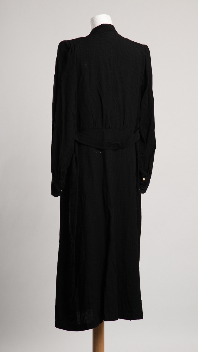 Fotsid svart damekjole med  krage. Åpning i overdel i front, hvit dekorflapp i åpningen, lukkes med trykknapper. I tillegg åpning i venstre sidesøm med trykknapplukking. Annet stoff innfelt i midjen og som panel i skjørtet i front. Dekorbelte i ryggen. To utenpå lommer i skjørtet i front. Skulderputer av vevet papir(antt). Bred manskjett  med dekorativ knappelukning, trukne øyeknapper.