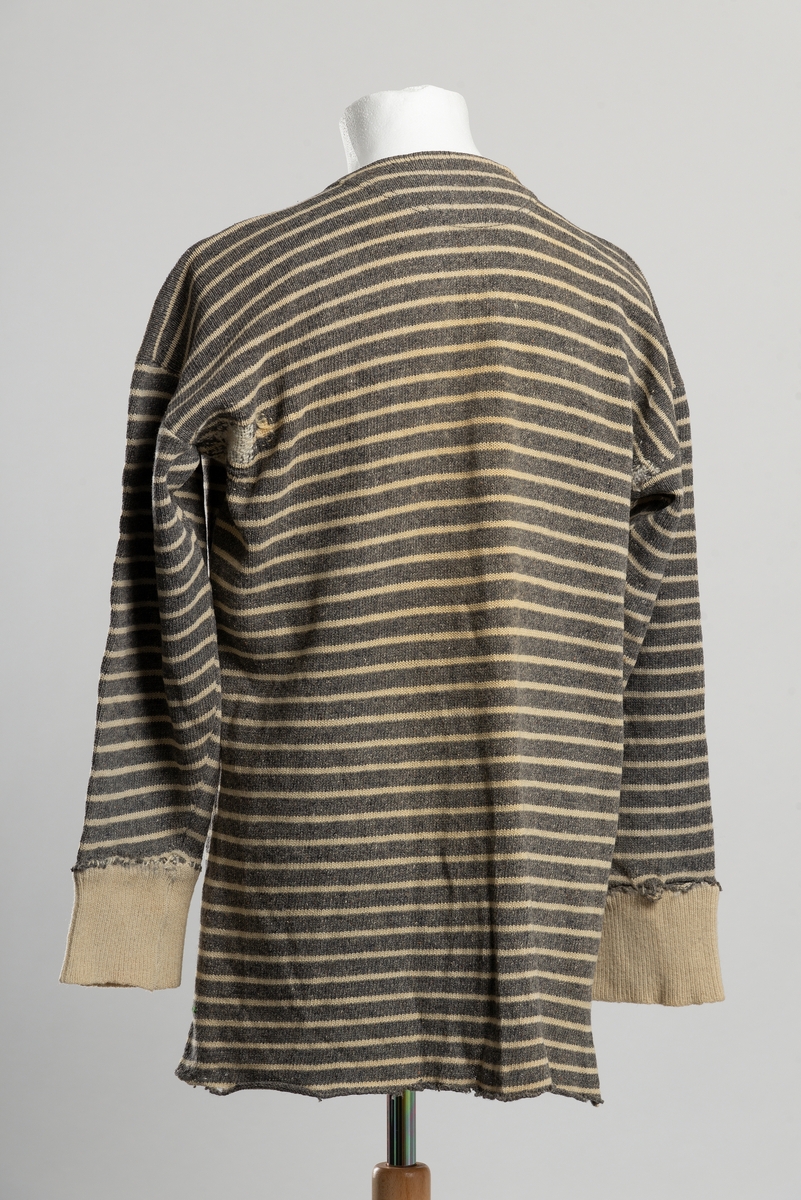 Grå strikket ullgenser med horisontale hvite striper. Åpning i front ned til brystet med knappelukkning, to perlemorsknapper. Manskjettene er nye å blitt sydd på i ettertid. Stoppinger ved skulder og under ermet. delvis heklet reparasjoni nederkant.