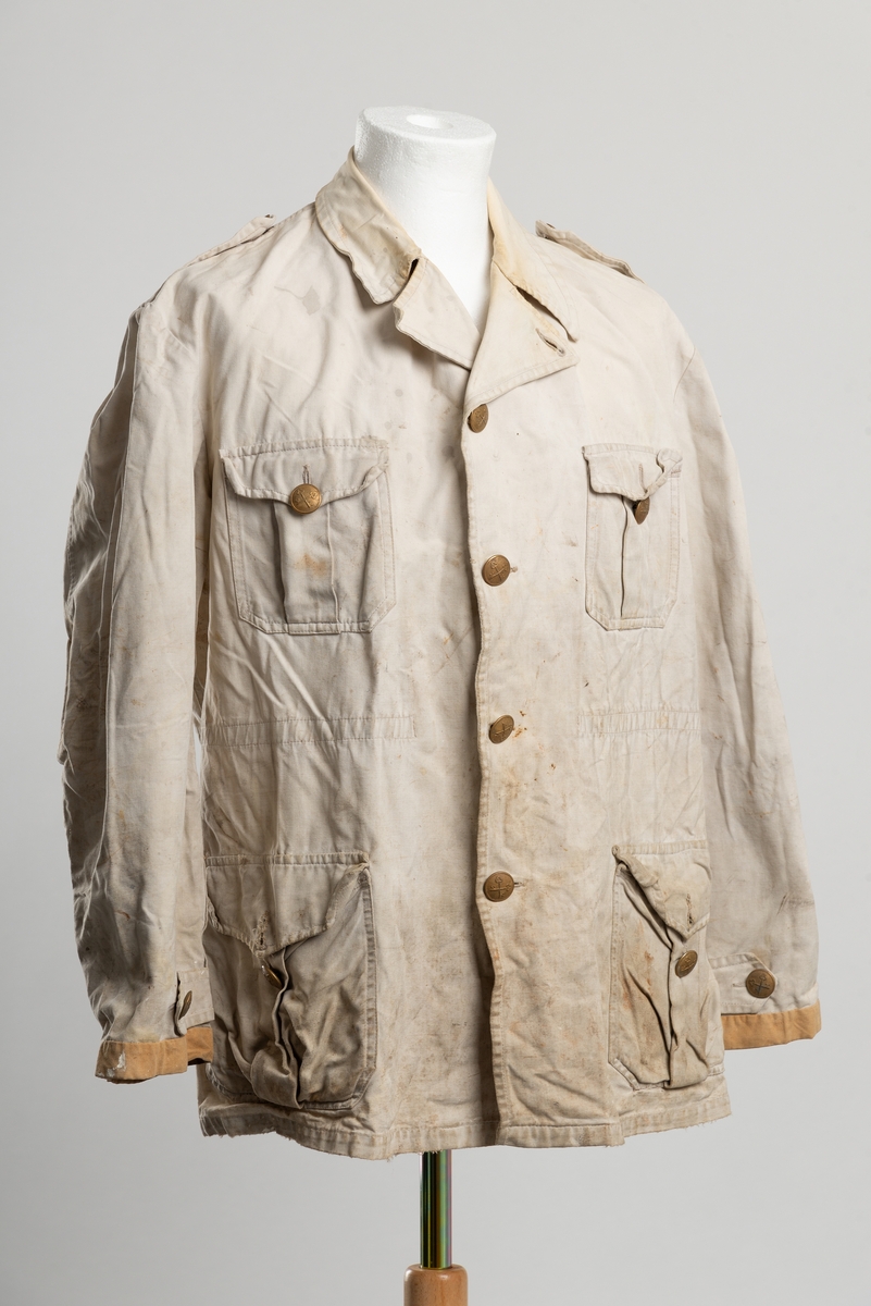 Arbeidsjakke av beige kanvas, med krage. Åpning i front med knappelukkning. Messingknapper med to korslagte nøkler som motiv..  Ekstra plastikknapp og knappehull ved halsen, slik at jakkeslaget kan brettes opp ved sterk vind. To brystlommer og to lommer i hoftehøyde med ekspansjon. Strikk i livet.  Epåletter med knapp på skuldre og nederst på erme. Nederst på ermet er lappet med ny kant.