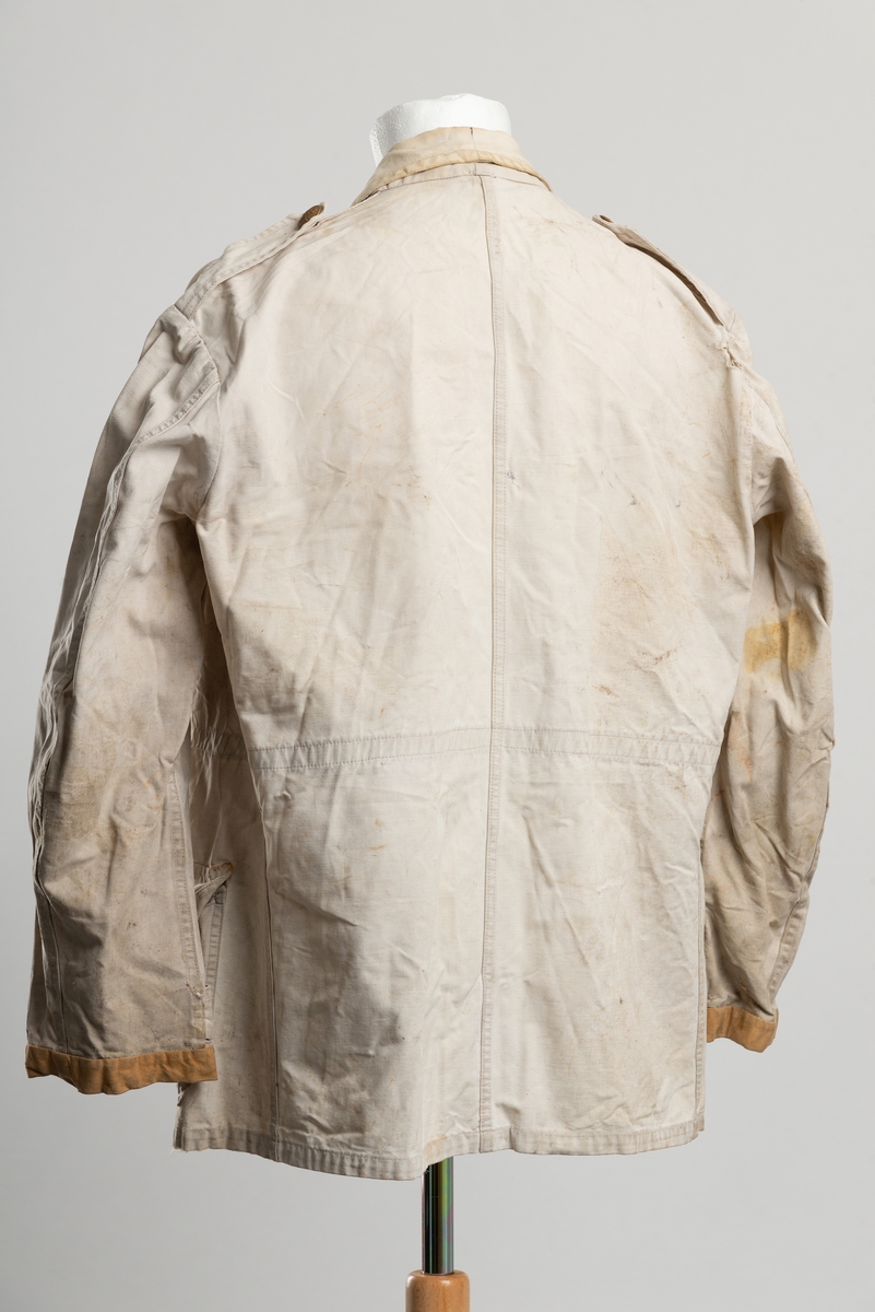 Arbeidsjakke av beige kanvas, med krage. Åpning i front med knappelukkning. Messingknapper med to korslagte nøkler som motiv..  Ekstra plastikknapp og knappehull ved halsen, slik at jakkeslaget kan brettes opp ved sterk vind. To brystlommer og to lommer i hoftehøyde med ekspansjon. Strikk i livet.  Epåletter med knapp på skuldre og nederst på erme. Nederst på ermet er lappet med ny kant.