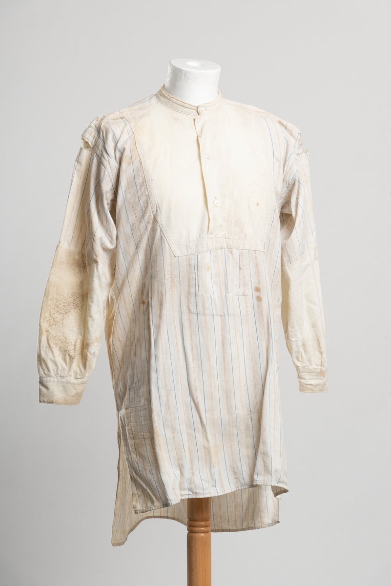 Lårlang herreskjorte med smale vertikale striper. Splitt i sidene ved hoftene. Bakstykket er lengre enn forstykket. Knapper av perlemor, antagelig håndlaget. Ermene er lappet og byttet ut med nye i ubleket lerret fra albuen i. Samme stoff er brukt til å lage ny hals/brystparti. Lappet i overgangen bryst-mage. Igjensydd hull ved høyre nederkant.