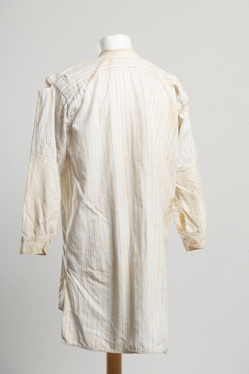 Lårlang herreskjorte med smale vertikale striper. Splitt i sidene ved hoftene. Bakstykket er lengre enn forstykket. Knapper av perlemor, antagelig håndlaget. Ermene er lappet og byttet ut med nye i ubleket lerret fra albuen i. Samme stoff er brukt til å lage ny hals/brystparti. Lappet i overgangen bryst-mage. Igjensydd hull ved høyre nederkant.