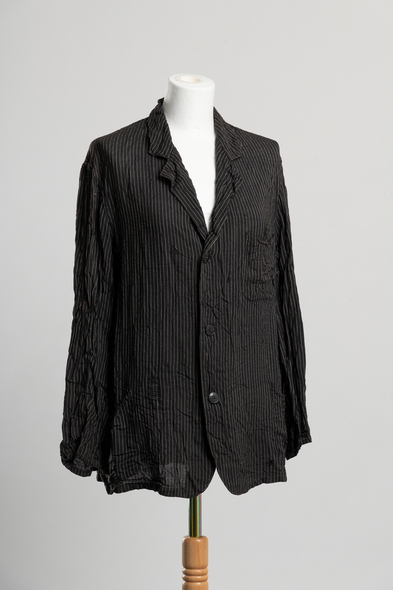 Jakke med krage og slag. Tynt stoff med vertikale smale striper. Åpning i front med knappelukkning, tre knapper. Brystlomme på venstre side og to lommer i hoftehøyde. Lappet ved lommer, i nakken og på ryggen.