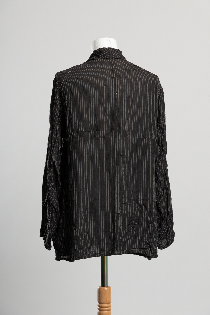 Jakke med krage og slag. Tynt stoff med vertikale smale striper. Åpning i front med knappelukkning, tre knapper. Brystlomme på venstre side og to lommer i hoftehøyde. Lappet ved lommer, i nakken og på ryggen.