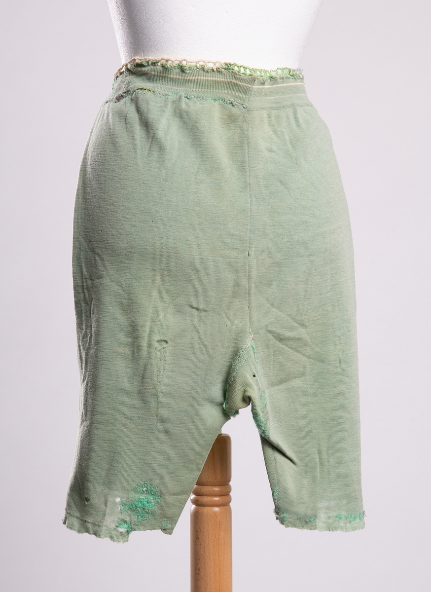 Lårlang dameunderbukse av bomullstrikot. Ribbekant i linningen. Diverse stoppinger ved linning, på lår, skrittet, og rumpe. Heklet kantreparasjon på linningen. Hull på venstre lår bak. Rester av produsent lapp i linningen.