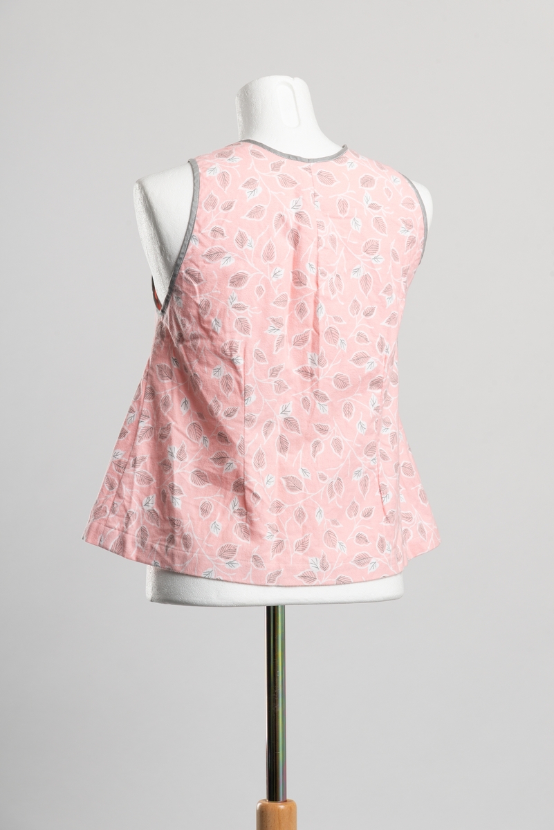 Hjemmesydd A-formet underliv av bomullsflanell med bladmønster. Ermehull og hals kantet med grått skråbånd. Åpning i front med knappelukkning, trukkede metallknapper.. To knapper langs falden i front.