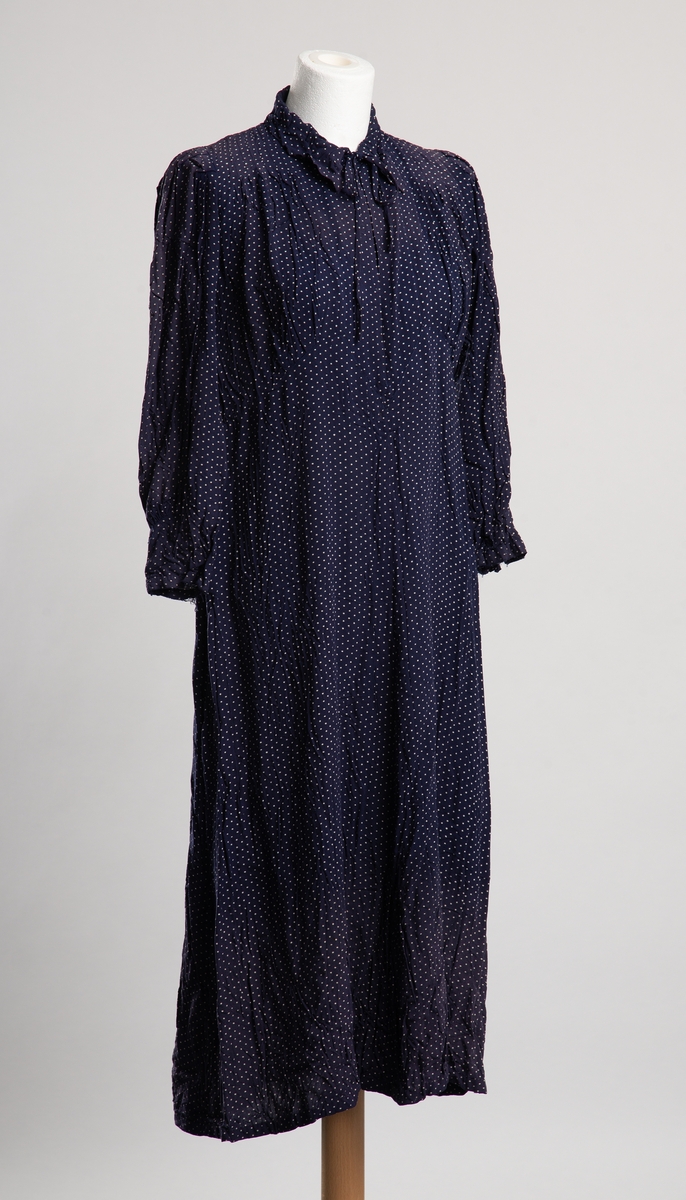 Knelang kjole av blått tekstil med hvite prikker. Krage med knytelukkning. Halssplitt med belegg med trykknapplukkning. Dekorative legg ved bryst. Sammenrynkede smale biskopermer.  Noe lappet i nakkeparti.