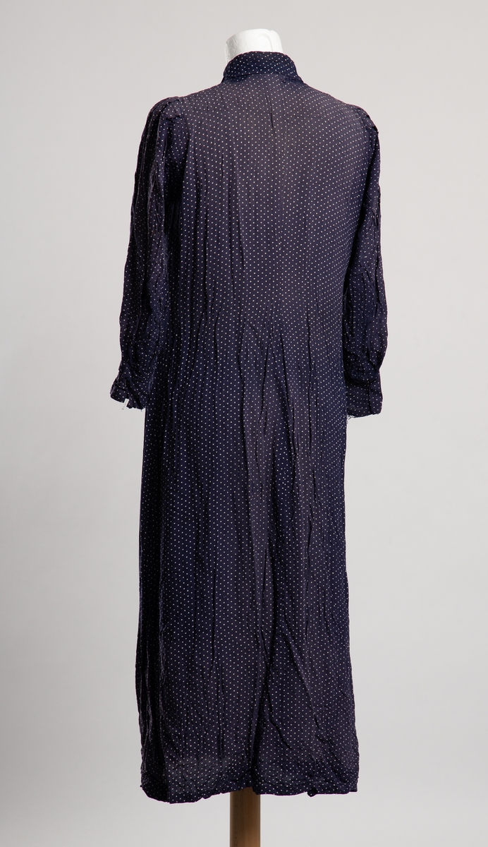 Knelang kjole av blått tekstil med hvite prikker. Krage med knytelukkning. Halssplitt med belegg med trykknapplukkning. Dekorative legg ved bryst. Sammenrynkede smale biskopermer.  Noe lappet i nakkeparti.