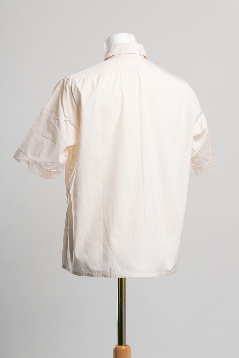 Kortermet skjorte med krage. Åpning i front med knappelukkning, fire plastknapper. Brystlomme på venstre side med klaff og knappelukning.