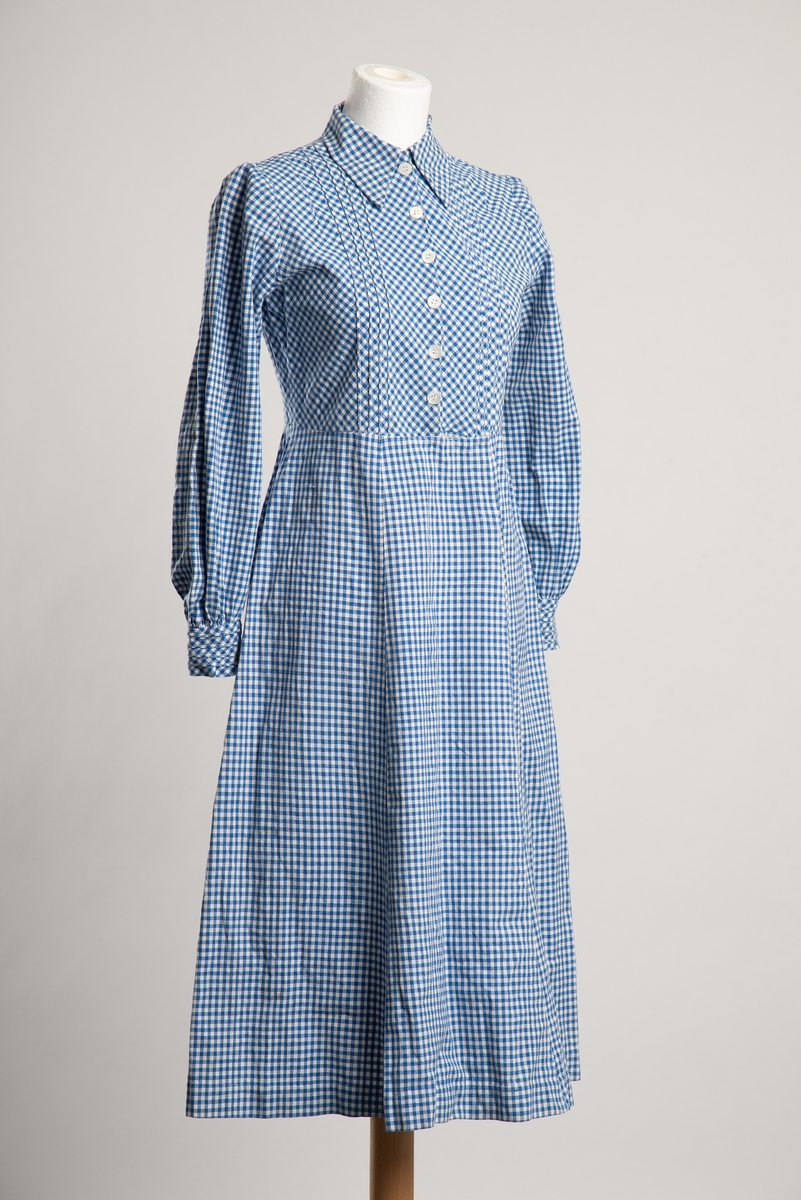 Legglang hvit og blå rutet kjole med krage. Åpning i front med trykknapplukning skult med dekorknapper utenpå. Dekorative bisesømmer på bryst og manskjetter.