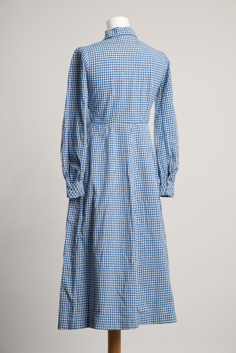 Legglang hvit og blå rutet kjole med krage. Åpning i front med trykknapplukning skult med dekorknapper utenpå. Dekorative bisesømmer på bryst og manskjetter.