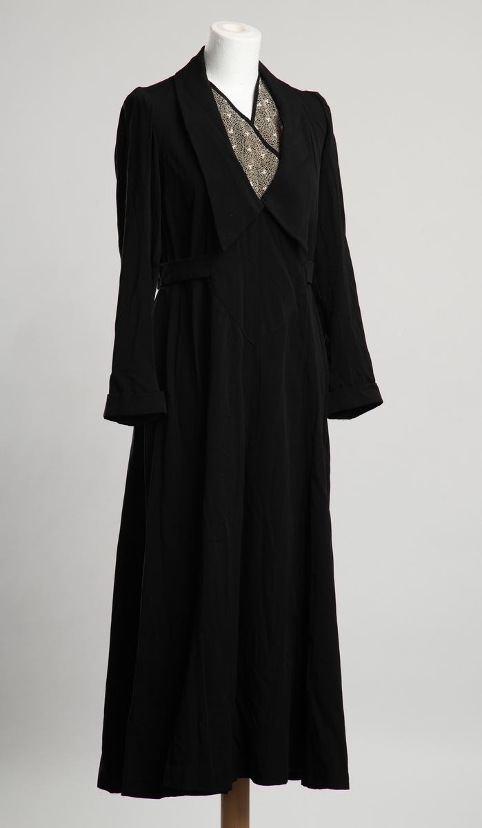 Svart fotsid damekjole. Krage med hvit snirkel- og blomsterbroderi halsåpningen. Åpning i front med trykknapplukkning. Belte i livet som festet i åpningen i front, festet i hemper i sidesømmene. Manskjetter med pynteknapp. Dekor på nederst på erme, to trekanter stikkende ut fra innsnitt nederst på erme,  i samme stoff som i halsåpningen. Antagelig hjemmesydd.