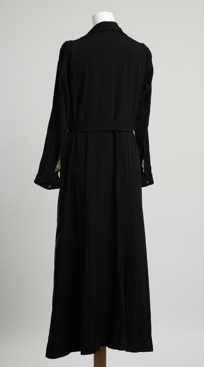 Svart fotsid damekjole. Krage med hvit snirkel- og blomsterbroderi halsåpningen. Åpning i front med trykknapplukkning. Belte i livet som festet i åpningen i front, festet i hemper i sidesømmene. Manskjetter med pynteknapp. Dekor på nederst på erme, to trekanter stikkende ut fra innsnitt nederst på erme,  i samme stoff som i halsåpningen. Antagelig hjemmesydd.