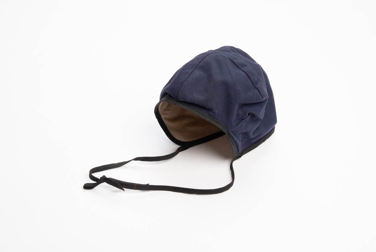 Blå lue av kypertvevet bomullstekstil, innvendig foret med gulhvit bomullsflanell. Kantet med svart skråbånd som ender i to snorer til å knytte under haka.