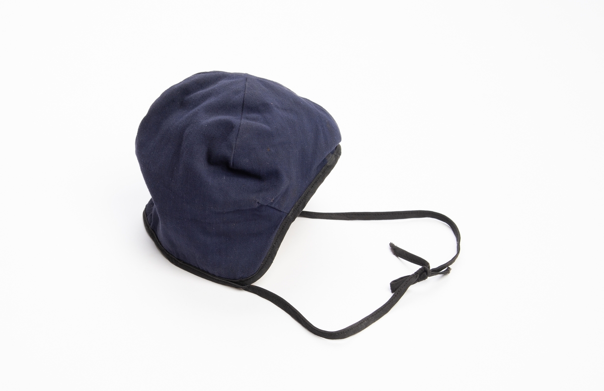 Blå lue av kypertvevet bomullstekstil, innvendig foret med gulhvit bomullsflanell. Kantet med svart skråbånd som ender i to snorer til å knytte under haka.